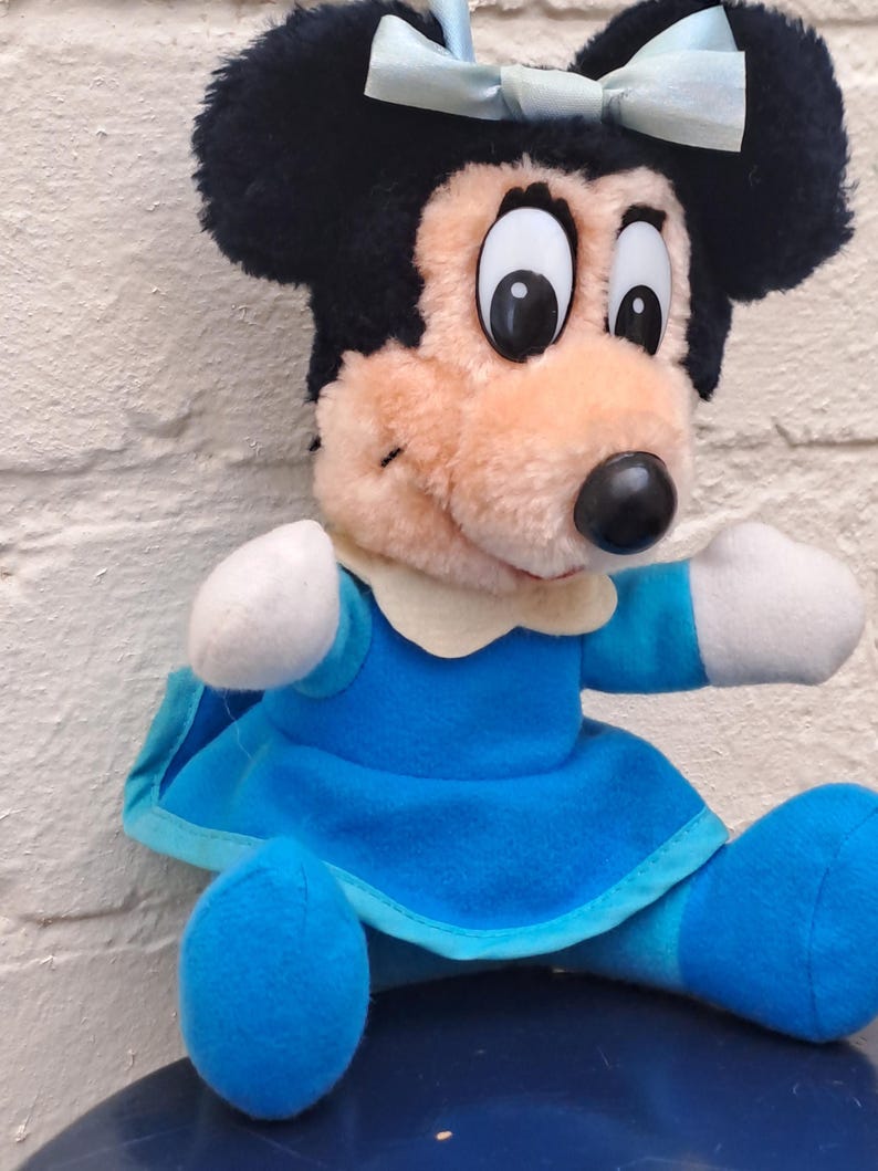 May include: A plush Minnie Mouse doll with black fur, large eyes, and a light blue bow. She wears a blue dress with white accents and blue shoes. The doll is sitting down.