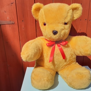Può includere: Un grande orsacchiotto di peluche giallo con un papillon rosso. L'orso è seduto su una superficie blu chiaro.