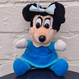 May include: A plush Minnie Mouse doll, featuring a black head with large ears and a light blue bow. The doll has a pink face, black eyes, and a black nose. She wears a blue dress with white accents and blue shoes. The doll is sitting.