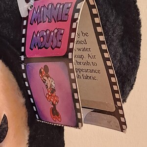 May include: A black and white plush Minnie Mouse toy with a pink tag that says "Minnie Mouse" in a bubble font. The tag also has a picture of Minnie Mouse in a red polka dot dress. The tag says "May be cleaned with water and soap. Air brush to appearance of plush fabric."