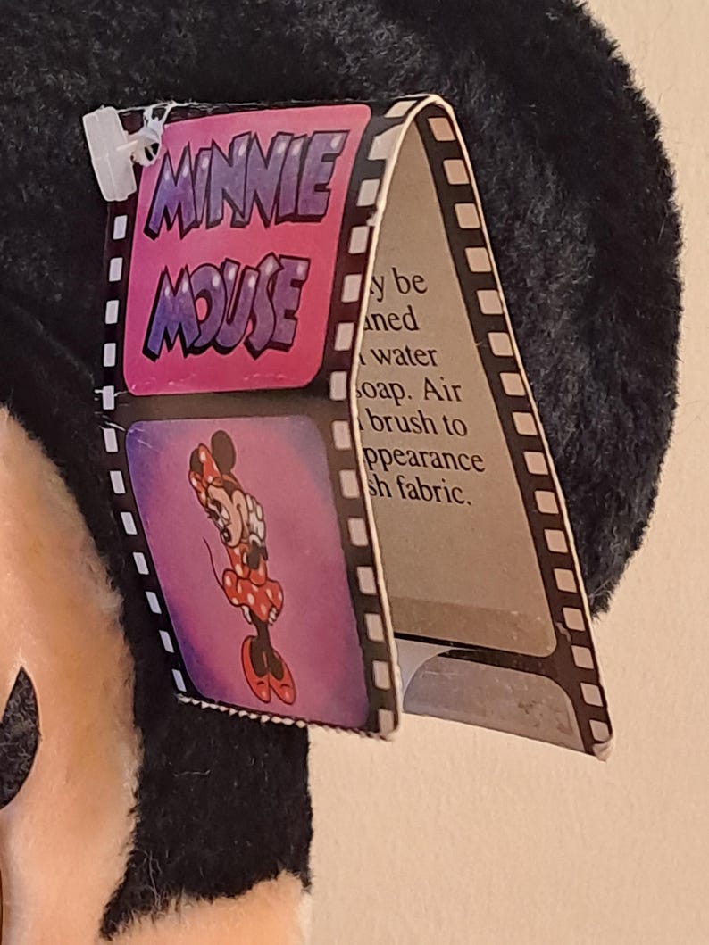 May include: A black and white plush Minnie Mouse toy with a pink tag that says "Minnie Mouse" in a bubble font. The tag also has a picture of Minnie Mouse in a red polka dot dress. The tag says "May be cleaned with water and soap. Air brush to appearance of plush fabric."
