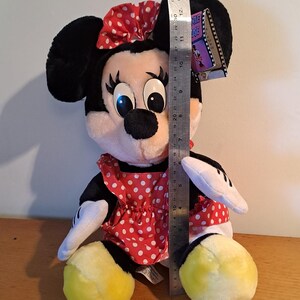 May include: A plush Minnie Mouse toy wearing a red dress with white polka dots and a red bow with white polka dots. The toy is sitting with its legs crossed and has yellow shoes. The toy is approximately 12 inches tall.