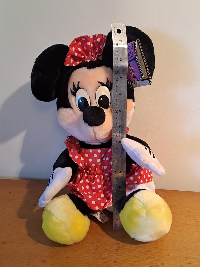 May include: A plush Minnie Mouse toy wearing a red dress with white polka dots and a red bow with white polka dots. The toy is sitting with its legs crossed and has yellow shoes. The toy is approximately 12 inches tall.
