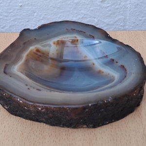 Könnte beinhalten: Eine polierte Agatschale mit natürlicher, organischer Form. Die Schale weist konzentrische Bänder in Grau-, Braun- und Blautönen auf, mit einem dunklen, strukturierten Außenrand. Das Innere hat eine glänzende, reflektierende Oberfläche.