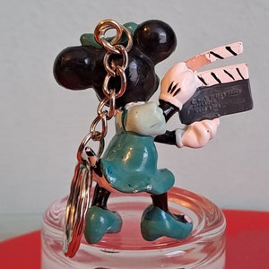 May include: A Minnie Mouse keychain figurine holding a film clapperboard. The figurine is black, white, and teal, with a gold-colored chain. The clapperboard has the text "Walt Disney Prod. Applause Hong Kong".