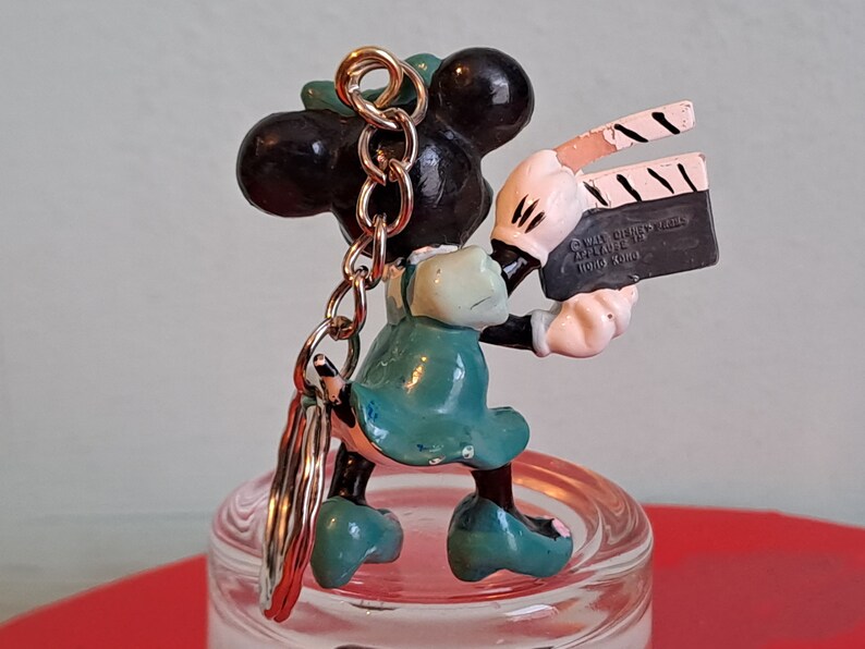 May include: A Minnie Mouse keychain figurine holding a film clapperboard. The figurine is black, white, and teal, with a gold-colored chain. The clapperboard has the text "Walt Disney Prod. Applause Hong Kong".