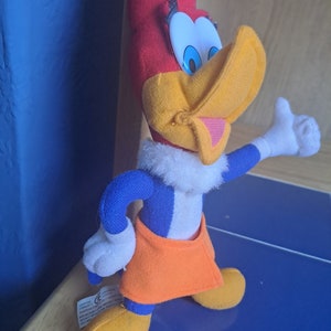 May include: A plush toy of Woody Woodpecker, a cartoon character. The toy is red, yellow, blue, and white. The toy is standing with its right arm raised and its right hand in a thumbs up gesture.