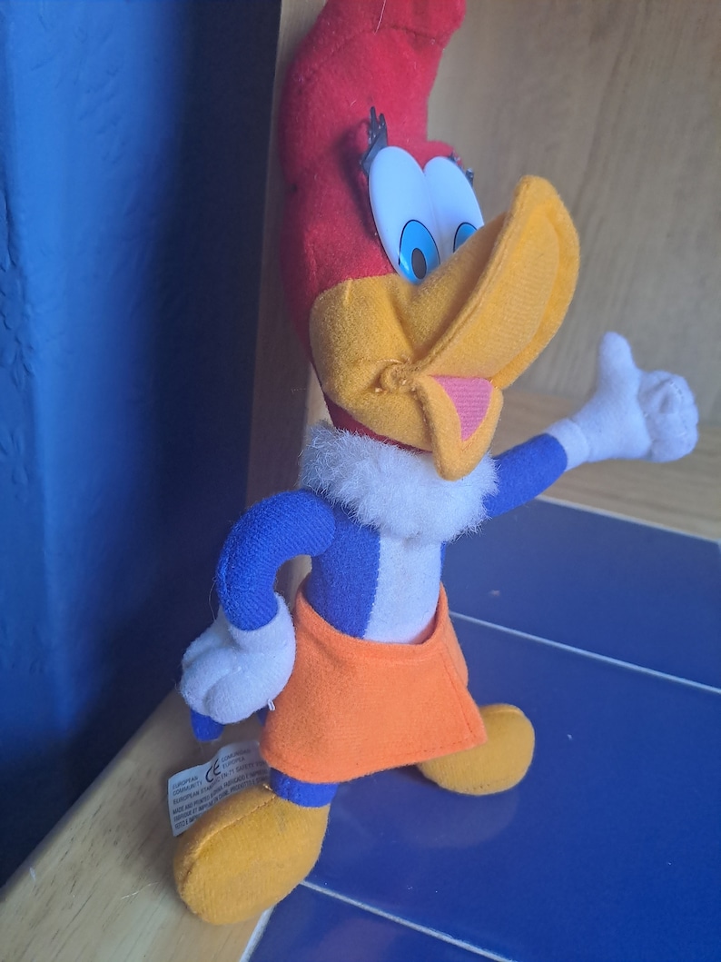 May include: A plush toy of Woody Woodpecker, a cartoon character. The toy is red, yellow, blue, and white. The toy is standing with its right arm raised and its right hand in a thumbs up gesture.
