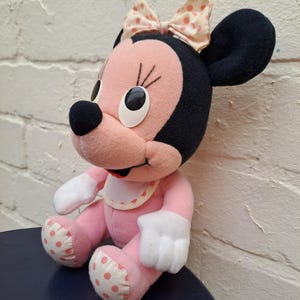 May include: A plush Minnie Mouse doll with a pink body, white gloves, and polka-dot accents. The doll has black ears, a pink face, and a bow with pink polka dots. The doll is sitting on a dark blue surface.