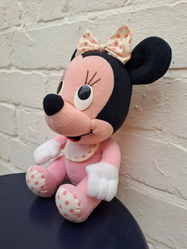 May include: A plush Minnie Mouse doll with a pink body, white gloves, and polka-dot accents. The doll has black ears, a pink face, and a bow with pink polka dots. The doll is sitting on a dark blue surface.