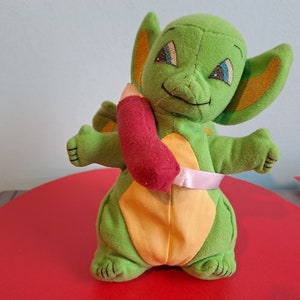 Peut inclure: Dragon en peluche vert avec des oreilles jaunes et orange, tenant un crayon en peluche rouge. Le dragon a un ruban rose autour de la taille.