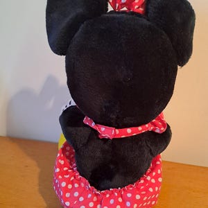 May include: Black plush toy with large ears and a red and white polka dot dress. The toy is sitting on a wooden surface.
