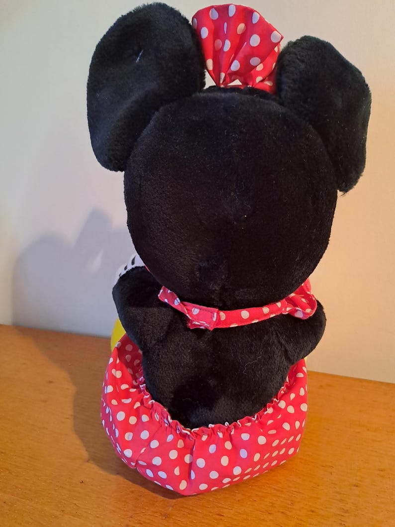 May include: Black plush toy with large ears and a red and white polka dot dress. The toy is sitting on a wooden surface.