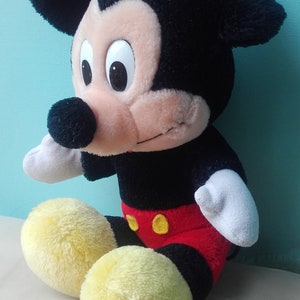 May include: A plush Mickey Mouse toy sitting with its legs crossed. The toy is black, red, and yellow with a big smile and large eyes.