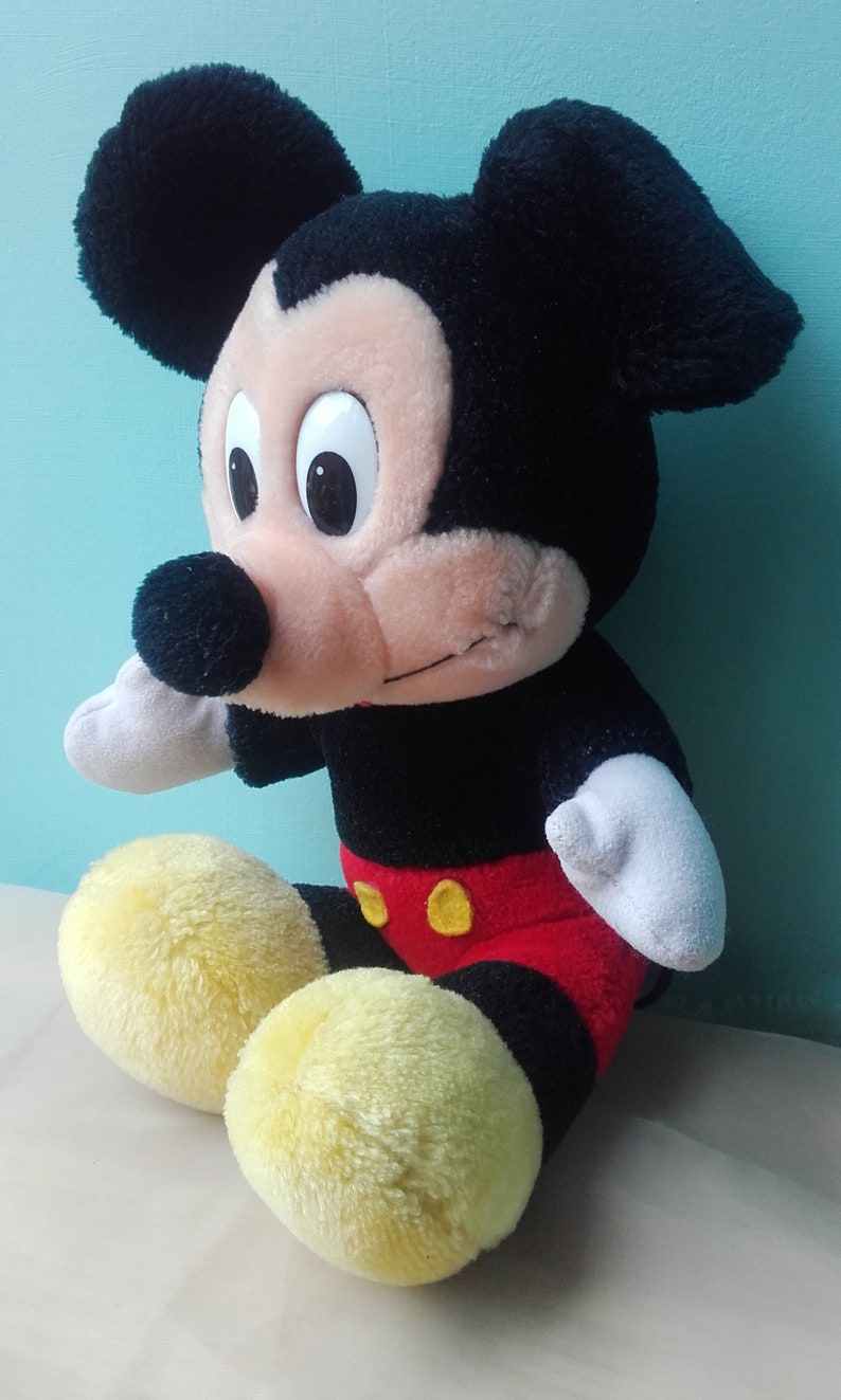 May include: A plush Mickey Mouse toy sitting with its legs crossed. The toy is black, red, and yellow with a big smile and large eyes.