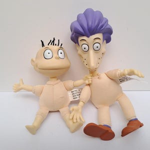 May include: Two character dolls with moulded plastic heads and soft fabric bodies. The smaller doll has a bald head with black hair tufts. The taller doll features spiky purple hair, a long nose, and brown shoes. Both dolls have light skin tones.