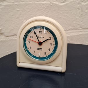 May include: A vintage cream-colored Westclox alarm clock. The clock face has a white background with black numerals and a teal outer ring. The clock's hands are black and red. The clock is on a dark blue surface.