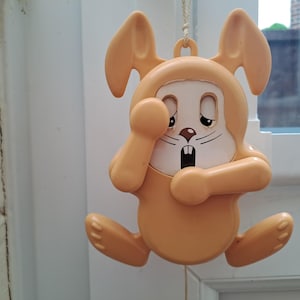 May include: A hanging orange bunny-shaped ornament with a sad expression. The bunny has large ears, a white face with black details, and is holding its paw up to its face. The ornament is made of plastic and is hanging from a white string.