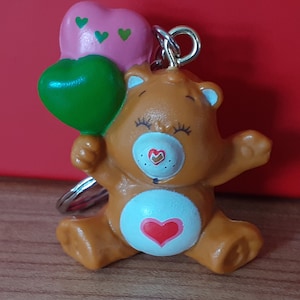 Może przedstawiać: Brelok w kształcie brązowego misia trzymającego różowe i zielone balony w kształcie serca. Miś ma biały brzuszek z czerwonym sercem i małym noskiem w kształcie serca. Brelok jest wykonany z błyszczącego materiału.