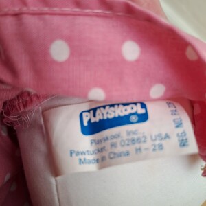May include: Close-up of a pink fabric with white polka dots. A tag with the Playskool logo and text is visible. The tag includes the company's name, address in Pawtucket, RI, and the text "Made in China".