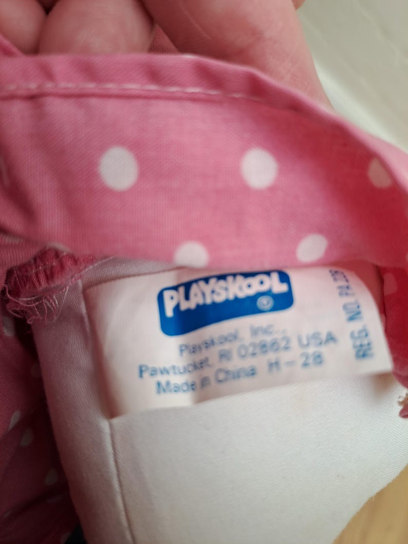 May include: Close-up of a pink fabric with white polka dots. A tag with the Playskool logo and text is visible. The tag includes the company's name, address in Pawtucket, RI, and the text "Made in China".