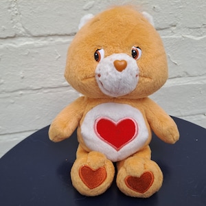Könnte beinhalten: Ein orangefarbener Care Bear-Plüschbär mit weißer Schnauze und einem roten Herz auf dem Bauch. Der Bär hat braune Augen und orangefarbene herzförmige Akzente auf den Füßen. Er sitzt auf einer dunkelblauen Oberfläche.