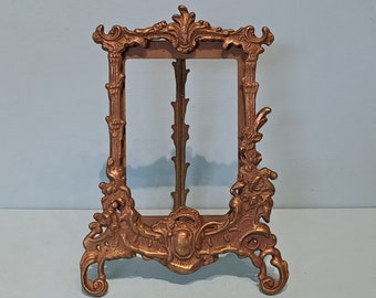 May include: A gold-toned metal picture frame with ornate details and a decorative base. The frame is designed to stand upright and has a rectangular opening for displaying a photo or artwork.