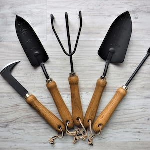 K&ouml;nnte beinhalten: F&uuml;nf Gartenwerkzeuge mit Holzgriffen und schwarzen Metallk&ouml;pfen. Die Werkzeuge umfassen eine Schaufel, einen Kultivator, eine Handharke, ein Unkrautwerkzeug und eine kleine Handschaufel.