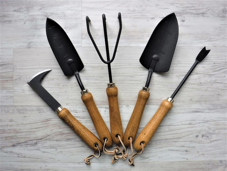 K&ouml;nnte beinhalten: F&uuml;nf Gartenwerkzeuge mit Holzgriffen und schwarzen Metallk&ouml;pfen. Die Werkzeuge umfassen eine Schaufel, einen Kultivator, eine Handharke, ein Unkrautwerkzeug und eine kleine Handschaufel.