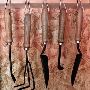 K&ouml;nnte beinhalten: F&uuml;nf verschiedene Gartenger&auml;te h&auml;ngen an einem Holzgestell. Die Ger&auml;te sind aus Metall und Holz und haben schwarze Griffe. Zu den Ger&auml;ten geh&ouml;ren eine Schaufel, ein Kultivator, eine Handharke, ein Unkrautstecher und eine kleine Schaufel.