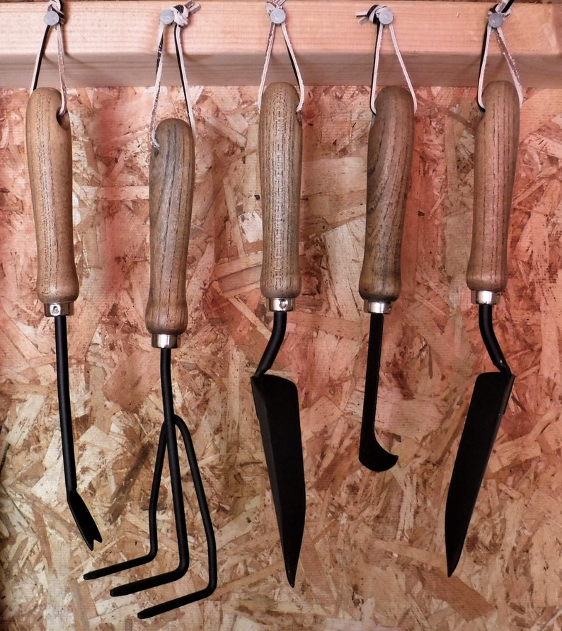 K&ouml;nnte beinhalten: F&uuml;nf verschiedene Gartenger&auml;te h&auml;ngen an einem Holzgestell. Die Ger&auml;te sind aus Metall und Holz und haben schwarze Griffe. Zu den Ger&auml;ten geh&ouml;ren eine Schaufel, ein Kultivator, eine Handharke, ein Unkrautstecher und eine kleine Schaufel.