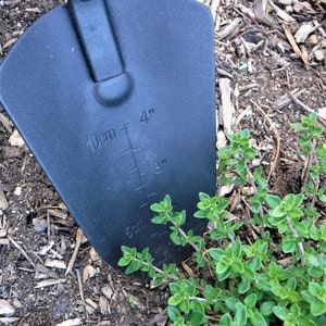 K&ouml;nnte beinhalten: Ein schwarzes Gartenwerkzeug mit einer 10 cm breiten Klinge und einer Messskala in Zoll und Zentimetern. Das Werkzeug liegt auf dem Boden neben einer gr&uuml;nen Pflanze.