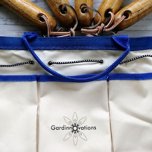 K&ouml;nnte beinhalten: Eine wei&szlig;e Gartenwerkzeugtasche aus Canvas mit blauem Besatz und einem schwarzen Kordelverschluss. Die Tasche hat mehrere Taschen und ein Logo mit der Aufschrift "Gardinnovations".