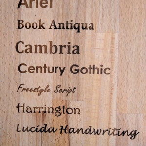 K&ouml;nnte beinhalten: Nahaufnahme einer Holzoberfl&auml;che mit einer Liste von Schriftarten f&uuml;r die Gravur. Die Optionen sind: Arial, Book Antiqua, Cambria, Century Gothic, Freestyle Script, Harrington und Lucida Handwriting.