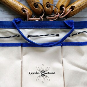K&ouml;nnte beinhalten: Eine wei&szlig;e Gartenwerkzeugtasche aus Canvas mit blauem Besatz und einem schwarzen Kordelverschluss. Die Tasche hat mehrere Taschen und ein Logo mit der Aufschrift "Gardinnovations".