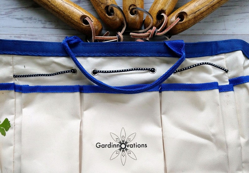K&ouml;nnte beinhalten: Eine wei&szlig;e Gartenwerkzeugtasche aus Canvas mit blauem Besatz und einem schwarzen Kordelverschluss. Die Tasche hat mehrere Taschen und ein Logo mit der Aufschrift "Gardinnovations".