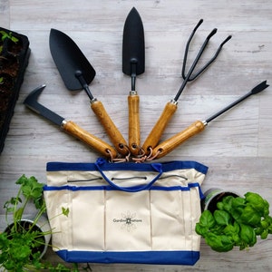 K&ouml;nnte beinhalten: Eine wei&szlig;e Gartenwerkzeugtasche aus Canvas mit blauem Besatz und dem aufgedruckten Text "Garden Innovations" auf der Vorderseite. Die Tasche ist gef&uuml;llt mit f&uuml;nf Gartenwerkzeugen: einer Schaufel, einem Handrechen, einem Kultivator, einer kleinen Schaufel und einem Handj&auml;te.