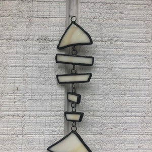 May include: A white stained glass fish bone hanging ornament with black outlines. The ornament is made of several pieces of glass that are connected by metal chains.
