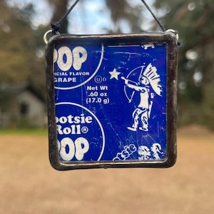May include: A square stained glass ornament featuring a vintage Tootsie Pop wrapper design. The blue background displays the text "POP" and "GRAPE", with an illustration of an archer. The net weight is .60 oz (17.0 g).