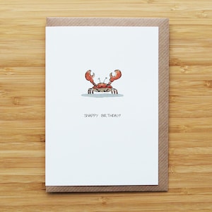 Peut inclure: Carte de vœux avec une illustration de crabe de dessin animé. Le crabe est rouge et marron avec de grandes pinces et est centré sur une carte blanche. Le texte "SNAPPY BIRTHAY!" est imprimé en dessous. La carte est dans une enveloppe marron.