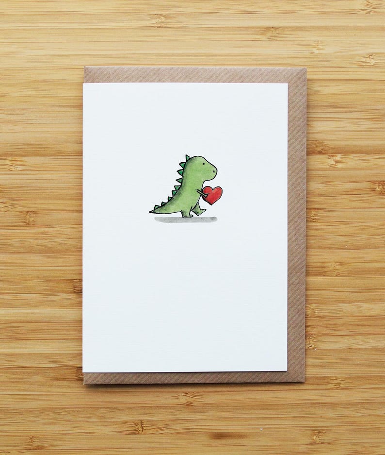 May include: A greeting card featuring a hand-drawn green dinosaur holding a red heart. The dinosaur is on a white card, set inside a brown envelope. The card is placed on a wooden surface.
