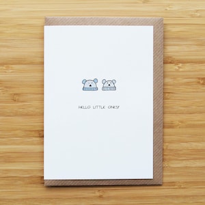 May include: A greeting card with a white front featuring two cartoon bear heads wearing blue hats. The text "HELLO LITTLE ONES!" is printed below the illustrations. The card is set inside a brown envelope, all on a wooden surface.
