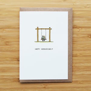 May include: A greeting card with a brown envelope. The white card features a hand-drawn illustration of a couple on a swing set, with the words "HAPPY ANNIVERSARY!" below the image. The card is set against a wooden background.