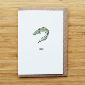 May include: A greeting card featuring a watercolor illustration of a green eel with a pink dorsal fin. The word "EELLO" is written in black below the eel. The card is on a brown envelope, set against a wooden background.