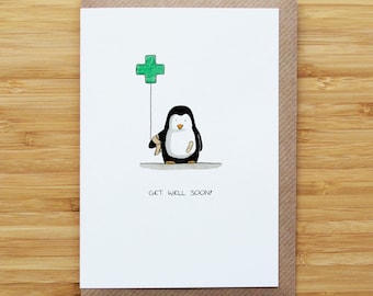 May include: A greeting card featuring a cartoon penguin with a bandage, holding a green cross balloon. The card has the words "GET WELL SOON!" printed below the penguin. The card is set on a wooden surface.