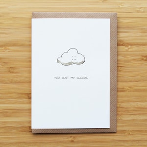Könnte beinhalten: Eine Grußkarte mit einer weißen Vorderseite mit einer Cartoon-Wolke mit einem lächelnden Gesicht. Der Text "YOU BUST MY CLOUDS." steht unter der Wolke. Die Karte befindet sich in einem braunen Umschlag auf einer Holzoberfläche.