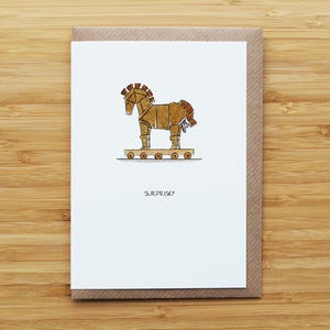 May include: A greetings card with a geometric illustration of a brown toy horse on wheels. The horse has a brown mane and tail. The word "SURPRISE!" is written below the horse. The card is set against a brown paper background.