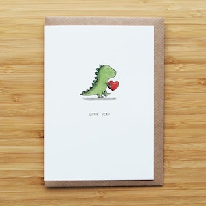 May include: A greeting card featuring a green dinosaur holding a red heart. The card has the words "LOVE YOU" printed below the dinosaur. The card is set on a brown envelope, with a wooden background.