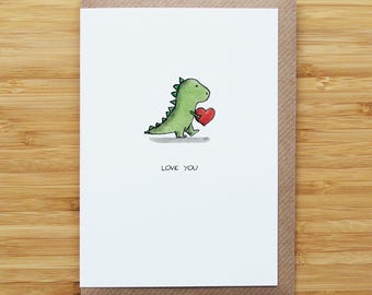 May include: A greeting card featuring a green dinosaur holding a red heart. The card has the words "LOVE YOU" printed below the dinosaur. The card is set on a brown envelope, with a wooden background.