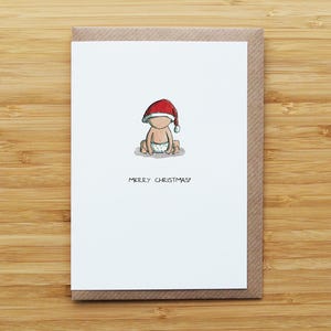 May include: A Christmas card with a cartoon baby wearing a Santa hat and a nappy. The baby is sitting down with the text "MERRY CHRISTMAS!" below the illustration. The card is on a brown envelope.